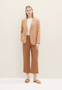 Light brown linen blazer with notch lapels, paired with a white sweater and brown wide-leg trousers. Brown slide sandals complete the look.