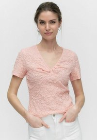 Lyserød T-shirt med korte ærmer og V-udskæring, tekstureret stof med et subtilt mønster og samlet detalje ved barmen. Hvide bukser med lommer.