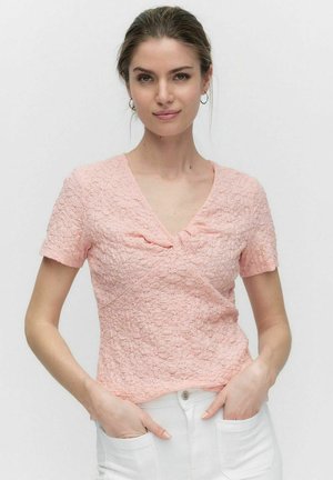 Rosa Kurzarmoberteil mit V-Ausschnitt, strukturiertem Stoff und dezentem Muster sowie Raffung an der Brust. Weiße Hose mit Taschen.