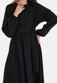 Mustassa pitkähihallisessa mekossa on pyöreä pääntie, rypistetyt hihansuut ja kankainen vyö vyötäröllä, sekä valuvat linjat.