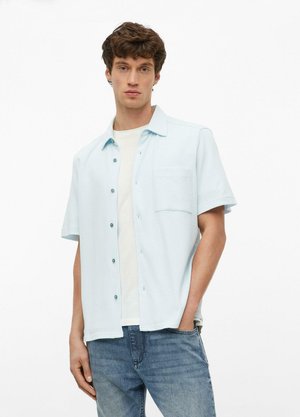 Junger Mann mit kurzen lockigen Haaren, trägt ein hellblaues kurzärmeliges Hemd über einem weißen T-Shirt und blaue Jeans, steht mit der Hand in der Hosentasche.
