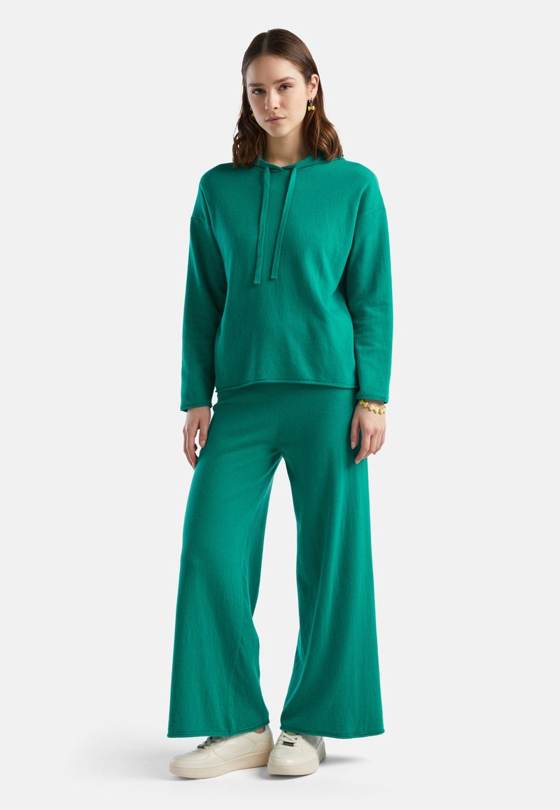 Sudadera con capucha color teal de corte relajado, combinada con pantalones de pierna ancha a juego. Ambos hechos de una tela suave. La modelo lleva zapatillas blancas.
