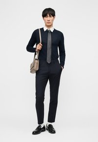 Jonge man die een donkerblauwe trui draagt over een wit overhemd met een grijze stropdas, zwarte broek, zwarte loafers, witte sokken en een taupekleurige schoudertas draagt.