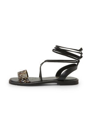 Sandalia negra con diseño de punta abierta, una tira con tachuelas en la parte frontal y cordones entrelazados que rodean el tobillo. Suela plana.