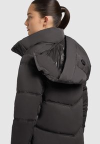 Sieviete ar tumšiem matiem zirgastē, tērpusies melnā pūkainā jakā ar lielu kapuci, redzama no muguras un sāniem uz gaiša fona.