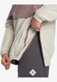 Ľahká polovičná zipsová bunda v krémovej a mauve farbe s elastickými manžetami, ktorá má bočné vrecká a zips. Šedé nohavice s logom.