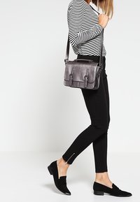 Grå läder crossbody-väska med två framfickor med spännen, justerbar axelrem och slät textur. Modell klädd i svarta åtsittande byxor och platta skor.