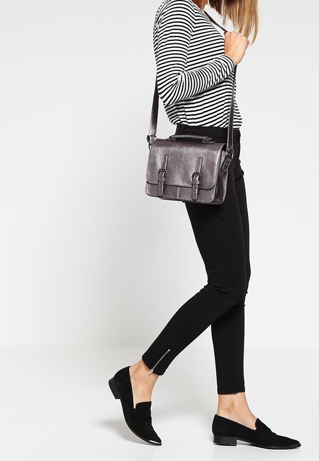 Grå läder crossbody-väska med två framfickor med spännen, justerbar axelrem och slät textur. Modell klädd i svarta åtsittande byxor och platta skor.