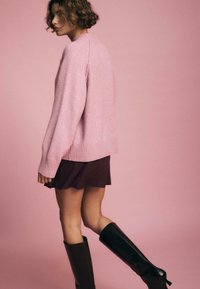 Helles rosa gestricktes Sweatshirt mit gerippten Bündchen und Saum, kombiniert mit einem dunkelbraunen Rock und knielangen schwarzen Stiefeln, vor einem rosa Hintergrund.