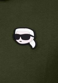 Grön stickad tyg med en tecknad ansiktsapplikation som har vitt hår och svarta solglasögon, omgiven av svart kontur.