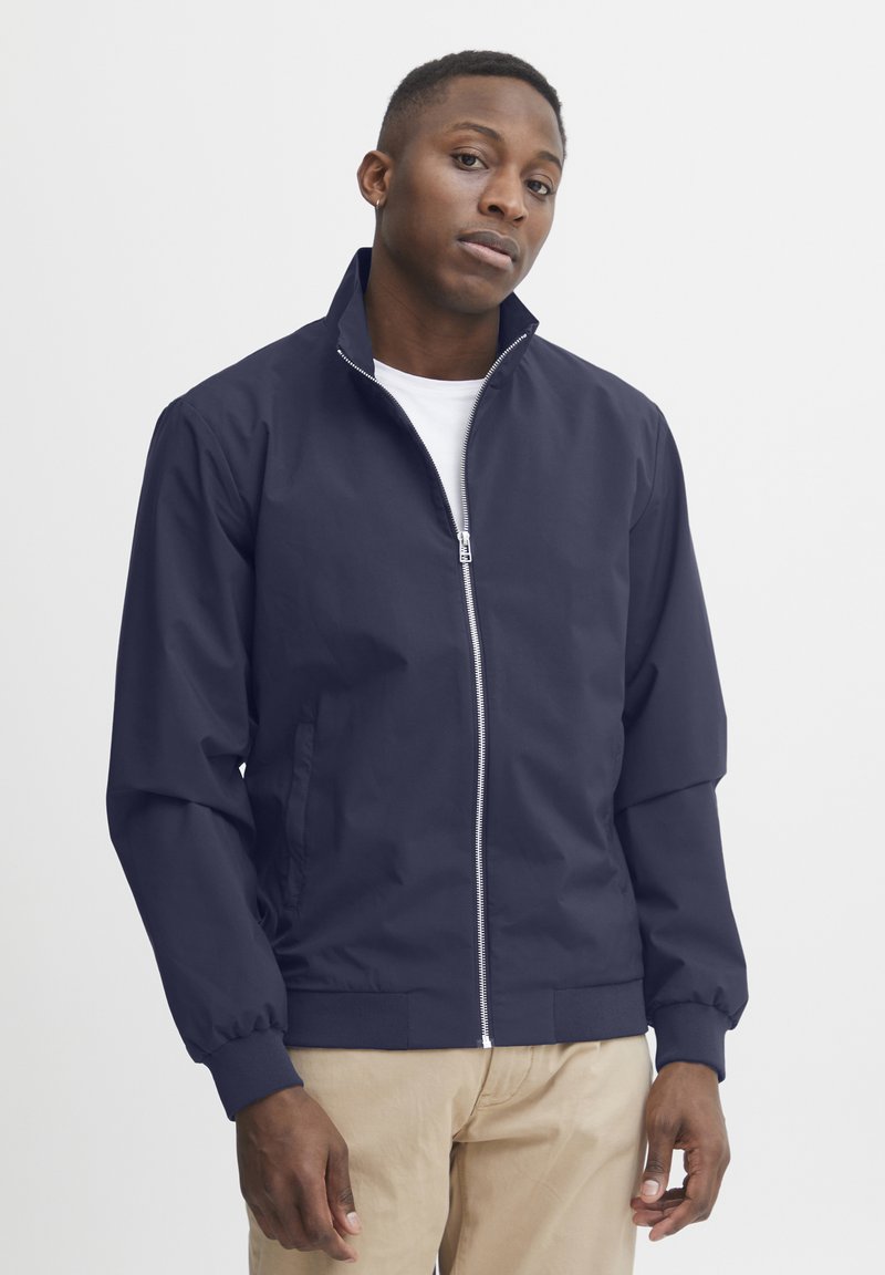 Marine windbreakerjack met ritssluiting, hoge kraag en elastische manchetten, gedragen over een wit shirt. Glad textuur met een minimalistisch ontwerp.