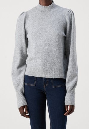 Personne portant un pull en tricot gris clair à manches longues avec un col rond et un jean bleu foncé ajusté.