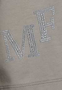 De letters "M" en "F" zijn gevormd met kleine zilveren rhinestones op een beige stof, met zichtbare stiksels aan de onderkant.