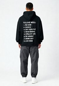 Uomo che indossa una felpa nera con cappuccio e testo bianco che elenca otto consigli di vita, pantaloni cargo scuri e sneakers bianche, rivolto di spalle su uno sfondo semplice.