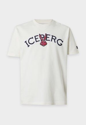 Мъж, облечен с бяла тениска Iceberg, тъмнокафяв панталон, червени маратонки, черна шапка, сребърно колие и гривни, стои с една ръка в джоба.