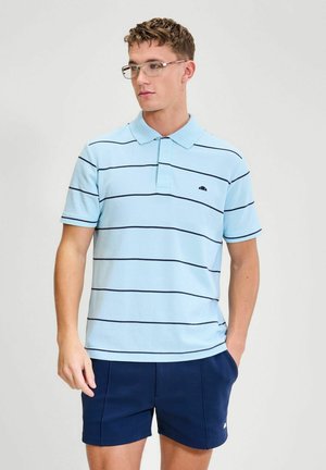 Młody mężczyzna w przezroczystych okularach, jasnoniebieskiej koszulce polo z ciemnymi poziomymi paskami i granatowych szortach, stoi z jedną ręką w kieszeni.