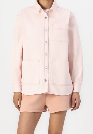 Persona que lleva una camisa de botones de color rosa pálido con bolsillos en el pecho y las caderas, combinada con pantalones cortos de color melocotón, mostrada desde el cuello hasta la mitad del muslo.