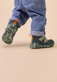 Scarpe per bambini in verde scuro con accenti grigi. Strappi in velcro, tomaia texture e suole a fantasia. Il modello indossa pantaloni in denim arrotolati.