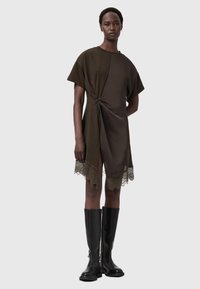 Olive zelené šaty s krátkymi rukávmi, detailom uzla na prednej strane a vlnitým čipkovým lemom. Nosené s čiernymi koženými čižmami po kolená. Hladká textúra.