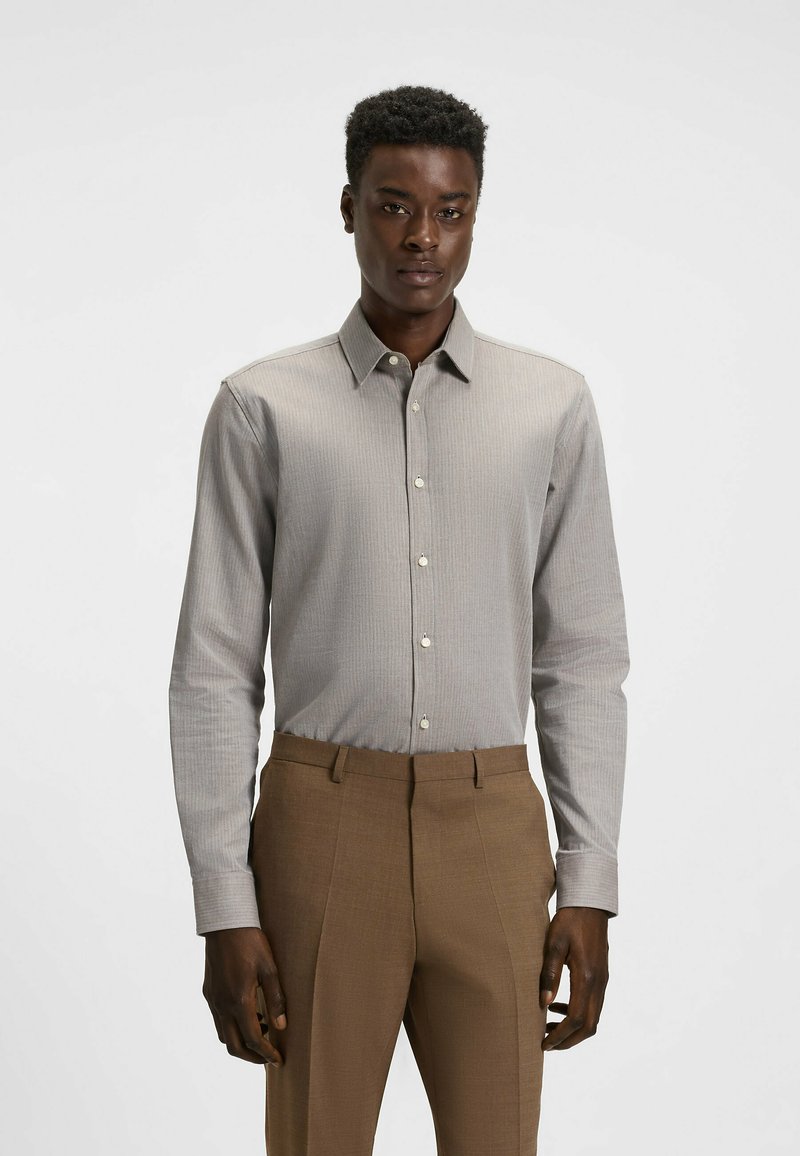 Camicia a righe grigio chiaro con colletto button-down e maniche lunghe, abbinata a pantaloni beige. Tessuto liscio con dettagli di texture sottili.
