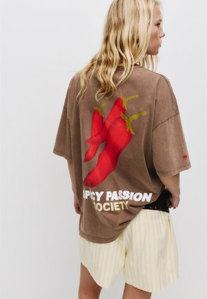 Brown oversized t-shirt featuring red chili pepper graphic and "SPICY PASSION SOCIETY" text. Soft material, relaxed fit, short sleeves.