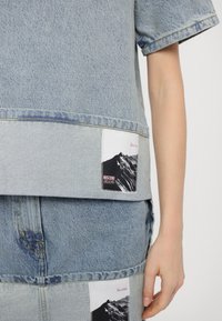 Džinsinė palaidinė su trumpomis rankovėmis, šviesiai mėlynos spalvos, su juosta apačioje. Puošia baltos dėmės su kalnų grafika ir "MOSCHINO JEANS" ženklu.