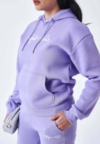 Felpa viola con cappuccio, tasca a marsupio, coulisse e logo ricamato sul petto. Abbinata a pantaloni coordinati e una borsa bianca con texture.