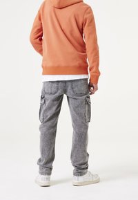 Person, der einen orangefarbenen Hoodie, graue Cargo-Jeans und weiße Nike-Sneaker trägt, steht mit entspannten Händen an den Seiten und schaut weg.