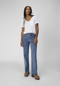 White cotton T-shirt with a pocket, paired with blue flared jeans. Model wears brown heeled clogs with silver accents. Plain background.