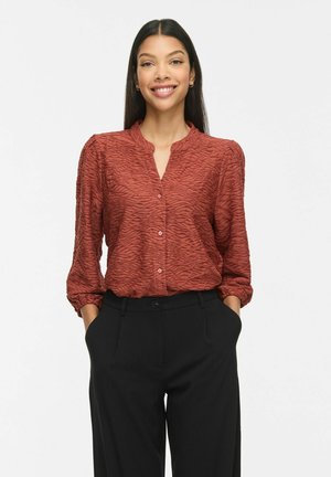 Femme aux longs cheveux foncés portant un chemisier texturé couleur rouille et un pantalon noir, souriante les mains dans les poches sur un fond blanc.