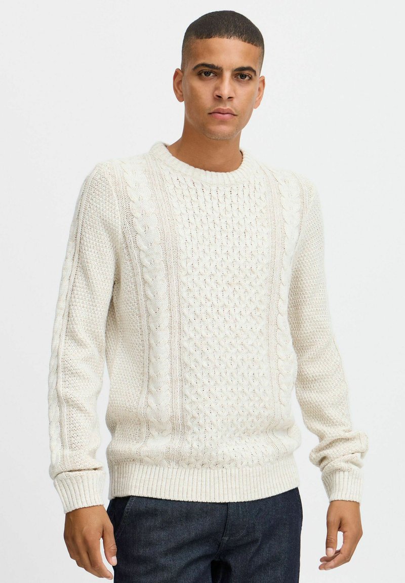 Suéter de punto color crema con un patrón de trenzas texturizadas, cuello y puños acanalados, y un corte relajado. Combinado con pantalones vaqueros oscuros.