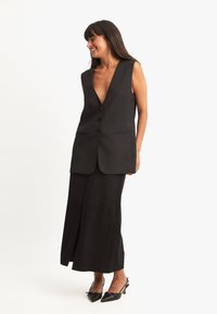 Must adornusega must ilma varrukateta vest sügava V-dekoltee, kahe esiküljel oleva tasku ja kolme nupuga, kombineerituna pika musta seeliku ja kontsadega. Sile tekstuur.