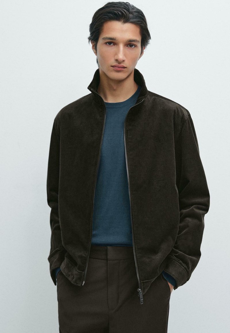 Brun kordflors zip-up jacka med hög krage, bärs över en blå rundhalsad tröja, kombinerad med mörka byxor. Subtil ribbad texturdetalj.