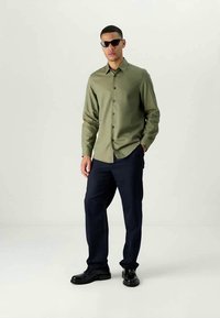 Άνδρας με σκούρα γυαλιά ηλίου, ελιάς πράσινη πουκαμίσα, ναυτικά παντελόνια και μαύρα παπούτσια, στέκεται μπροστά σε απλό ανοιχτό φόντο.