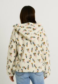 Veste en polaire à capuche crème avec des motifs floraux en rose, marron et vert, présentant une texture douce et une coupe décontractée.