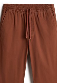Pantalons de jogging couleur rouille avec une taille élastique et un cordon de serrage, dotés de poches latérales et d'une texture de tissu lisse et léger.