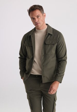 Uomo che indossa giacca e pantaloni verde oliva con maglione beige, in piedi davanti a uno sfondo semplice grigio chiaro.