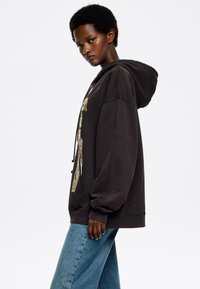 Sudadera negra de gran tamaño con estampado gráfico dorado, capucha con cordón y puños acanalados, combinada con jeans de mezclilla azules. Textura suave, corte relajado.