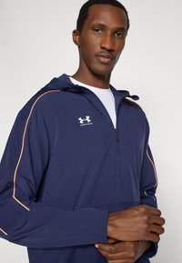 Ελαφριά μπλε ναυτικού χρώματος ζακέτα με μισό φερμουάρ, σχεδιασμό με κουκούλα, πορτοκαλί λεπτομέρειες κατά μήκος των μανικιών και μικρό λευκό λογότυπο στο στήθος.