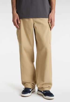 Beige cargobroek met wijde benen, voorzien van twee zijzakken. Draagt een donkergrijze shirt en marineblauwe sneakers met witte accenten.