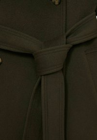 Olivroheline villane mantel lähivõtt, millel on seotud vöö ja suured nööbid, pehme ja tekstureeritud kangas. Näha on õmbluse detaile.