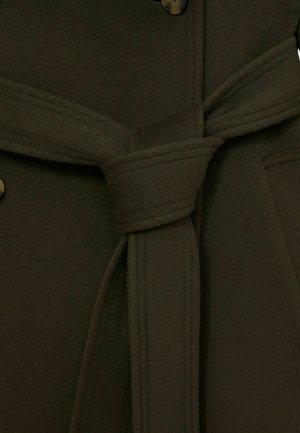 Cămașă din lână verde măslin, cu detalii în prim-plan, având un cordon la talie, nasturi mari și un material moale, texturat. Detalii vizibile ale cusăturii.