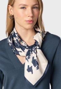 Zijden sjaal met een crèmekleurige basis en abstracte marineblauwe patronen. Zachte textuur, vierkante vorm, gedragen geknoopt om de hals over een donkere top.