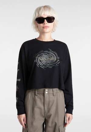 Maglietta nera a maniche lunghe e taglio cropped con un logo Vans grigio a spirale sul davanti. Il modello indossa occhiali da sole neri oversize e pantaloni cargo beige.