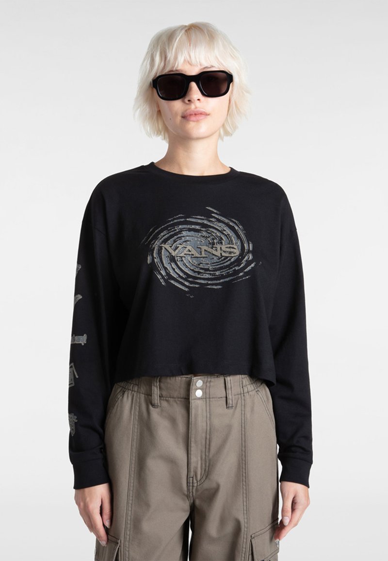 Zwarte cropped long-sleeve shirt met een grijze spiraal Vans-logo op de voorkant. Het model draagt oversized zwarte zonnebril en beige cargobroek.