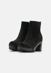 Un par de botas de tacón ancho de ante negro con detalle de tachuelas en un fondo blanco.