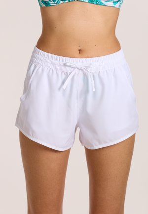 Frau trägt weiße Shorts mit elastischem Bund und Kordelzug sowie ein grünes, gemustertes, kürzer geschnittenes Oberteil, das den Bauch freilegt, vor einem schlichten Hintergrund.