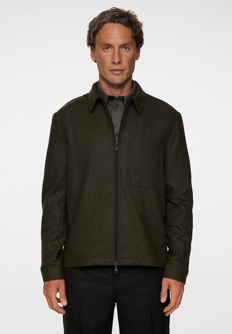 Mörkgrön jacka med dragkedja gjord av texturerad tyg, med spetsig krage och en bröstficka. Skräddarsydd passform med långa ärmar.