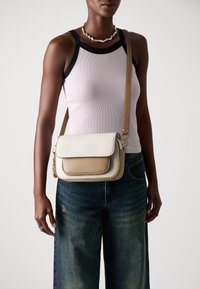 Crossbody-veske i beige og lys brun teksturert skinn, med klaffevern og detaljer i gull. Bærer med casual blå jeans.