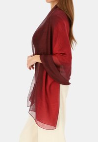 Przezroczysty, prostokątny szal w gradiencie od ciemnej czerwieni do jaśniejszego koloru burgunda. Lekka tkanina miękko opada, z płynnymi rękawami i bez zapięć.